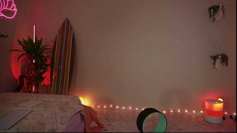 Media: Video of a dimly lit bedroom with beige walls, a green plant, surfboard leaning against the wall, string lights, and a yoga mat with a green circle on the floor.