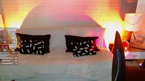 Media: A video of a modern, minimalist bedroom with a white bed, black pillows with white geometric patterns, and a colorful, abstract light display on the wall.