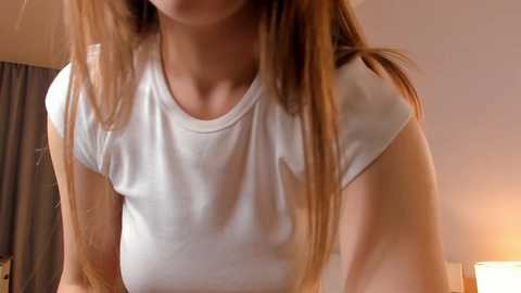 Media: Video of a young woman with light skin and long, straight brown hair, wearing a white, short-sleeved t-shirt that clings to her petite, slender frame, revealing subtle contours. Background shows a dimly lit room with a lamp and curtains.