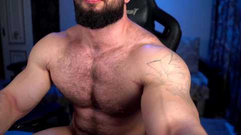 Media: Video of a shirtless, muscular man with a beard, hairy chest, and tattoos, sitting in a gaming chair in a dimly lit room with blue lighting.