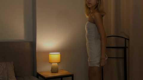 Media: Video of a slender, blonde woman in a strapless white dress, standing in a dimly lit bedroom with a wooden nightstand featuring a lamp, and a wardrobe visible in the background.