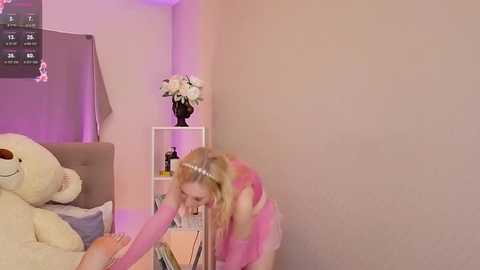 Media: Video of a young woman with blonde hair in a pink wig, kneeling beside a child in a white nightgown, in a soft-lit, pastel-colored bedroom with a teddy bear and floral decor.