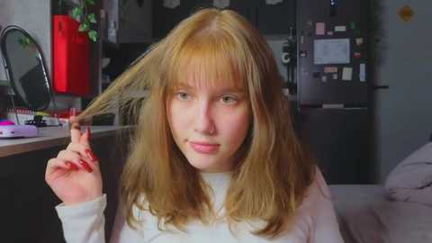 Media: Video of a young woman with long, straight blonde hair, holding a section with one hand, wearing a white long-sleeve top. Background features a cluttered kitchen with a red cabinet, a round mirror, and a black refrigerator.