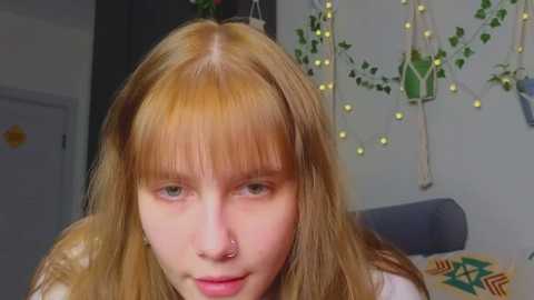 Media: Video of a young woman with long, straight, light brown hair and pale skin, wearing a white shirt. Background features a gray couch, a wall with green hanging plants, and yellow string lights.