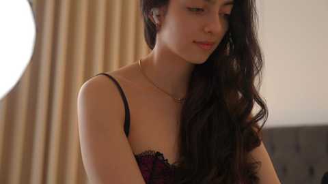 Media: Video of a young woman with long, dark wavy hair, wearing a black lace bra, standing indoors near beige curtains.