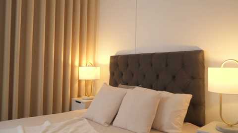 Media: Video of a modern, minimalist bedroom featuring a tufted, dark gray headboard, beige bedding, and two side tables with matching lamps casting a warm glow. The background includes floor-length beige curtains.