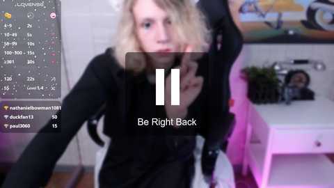 Media: A blurry video of a blonde woman in a black outfit, holding a phone, in a room with a pink light, a white desk, and a poster in the background. Text overlay says \"Be Right Back.\