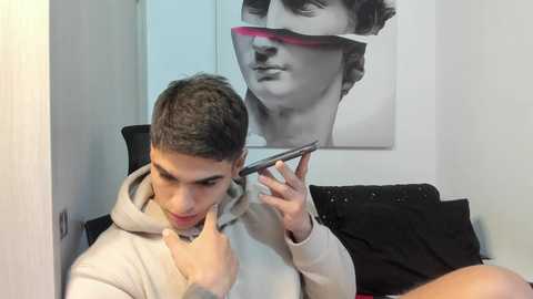 Media: Video of a young man with short dark hair, wearing a light-colored hoodie, adjusting his smartphone's camera. Behind him, a large black-and-white poster of a man's face.