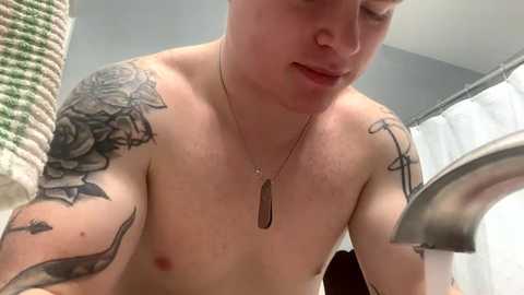 Media: Video of a shirtless, fair-skinned person with tattoos, including a large rose on their left shoulder, holding a metal shower head in a bathroom with a white curtain and green towel.