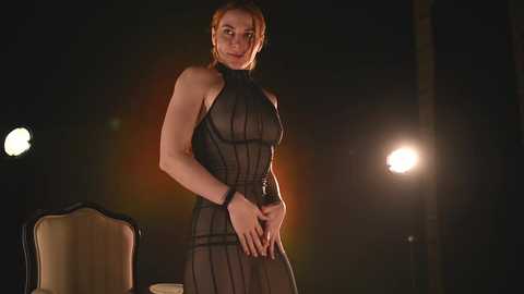 Media: Video of a fair-skinned, red-haired woman with medium build, wearing a sheer black halter dress with a plunging neckline, standing against a dimly lit background with a spotlight, hinting at a dramatic or theatrical setting.