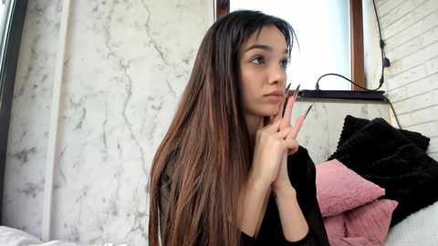 Media: Video of a young woman with long, straight, dark hair, light skin, and brown eyes, holding a manicure stick, in a cozy bedroom with white walls, a brick wall, pink and black pillows, and a window.