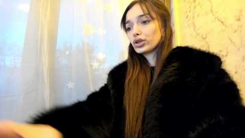 Media: Video of a young Caucasian woman with long brown hair, wearing a black fur coat, standing in front of a semi-transparent white curtain with a blurred cityscape background.