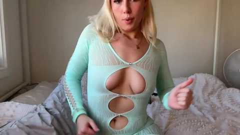 Media: Video of a blonde woman with fair skin wearing a revealing, mint-green, long-sleeve bodysuit with cutouts, revealing ample cleavage, on a bed in a dimly lit room.