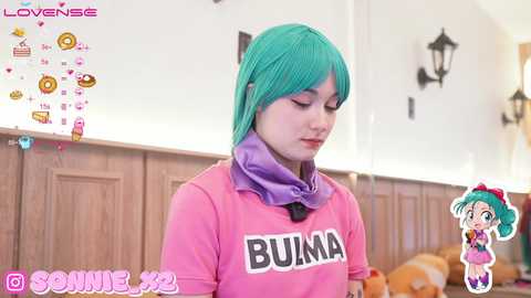 Media: A video of a woman with teal hair in a pink \"BUNA\" sweatshirt, wearing a purple scarf, in a cozy, wooden-paneled room. The background features a black wall sconce and a cartoon character.