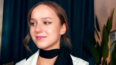 Media: Video of a young woman with light skin and brown hair styled in loose waves. She wears a black choker and a white jacket, with a teal curtain backdrop and a green plant in the background.