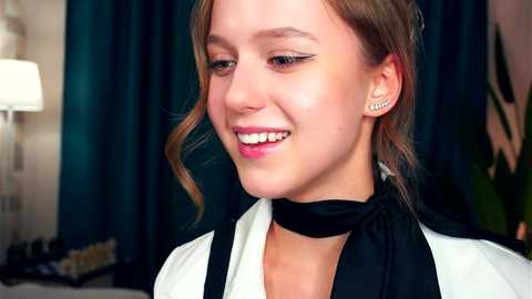 Media: Video of a young woman with light brown hair in a side ponytail, smiling, wearing a black choker and white blouse with black trim. Background features dark blue curtains and a green plant.