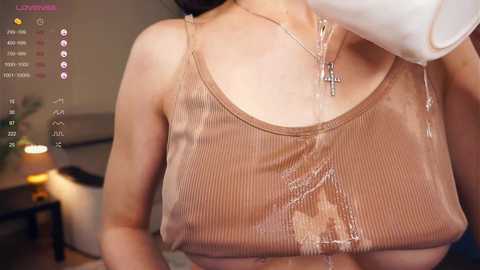 Media: A video of a woman in a wet, beige ribbed tank top, revealing her nipples through the fabric, wearing a cross necklace. Background shows a dimly lit room with a lamp and blurred furniture.