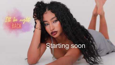 Media: Video of a light-skinned woman with long, curly black hair, lying on her stomach in a gray dress. Text reads \"I'll be right back\" and \"Starting soon.\