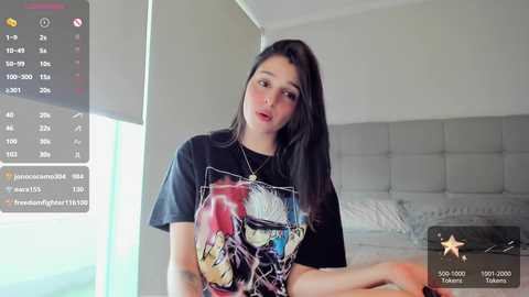 Media: Video of a young woman with long black hair, wearing a graphic T-shirt, sitting on a bed in a bright, modern bedroom.