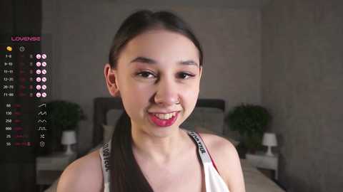 Media: Video of a young East Asian woman with long black hair, light skin, and pink lipstick, smiling in a modern bedroom with a white bed, green potted plants, and a gray wall.