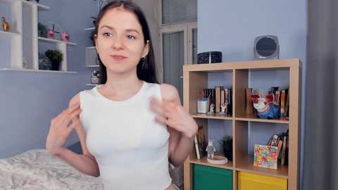 Media: A video of a young, slim woman with straight dark hair, wearing a white crop top, standing in a light blue room with a wooden shelf filled with books and toys.