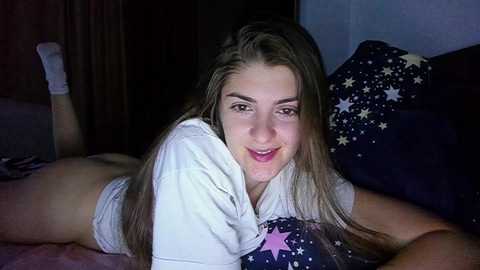 Media: Video of a young Caucasian woman with long brown hair, lying on a bed, wearing a white T-shirt and gray underwear, smiling. Background features a dark wooden headboard and a pillow with starry-patterned fabric.