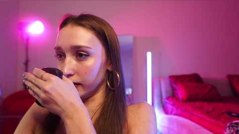 Media: Video of a young woman with long brown hair, wearing large hoop earrings, holding a cigarette close to her face, in a room with red walls and bed, under pink lighting.