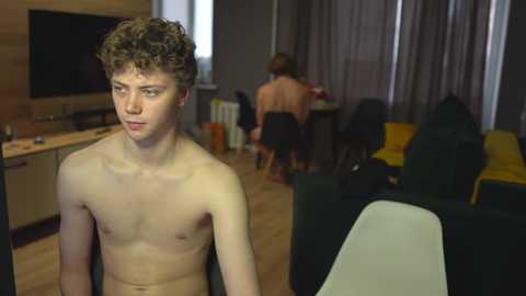 Media: Video of a young, shirtless, curly-haired, fair-skinned man in a modern living room with beige walls and wooden floors. Background shows a woman in a black hoodie and another man in a black shirt.