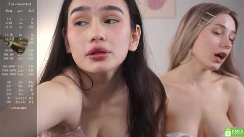 Media: Video of two young Asian women with long hair, one with dark hair and a pink bra, the other blonde with a hairclip, both topless. Background includes a calendar and abstract art.