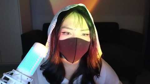 Media: Video of an East Asian woman with straight, shoulder-length hair, wearing a white hoodie and a black face mask, holding a glowing blue handheld device. The background is dimly lit with warm tones.