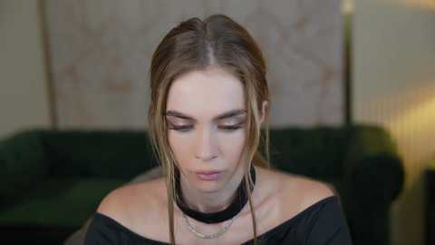 Media: Video of a fair-skinned, blonde-haired woman with a black choker, wearing a black off-shoulder top, looking downward, in a dimly-lit room with a green couch and abstract wall art in the background.