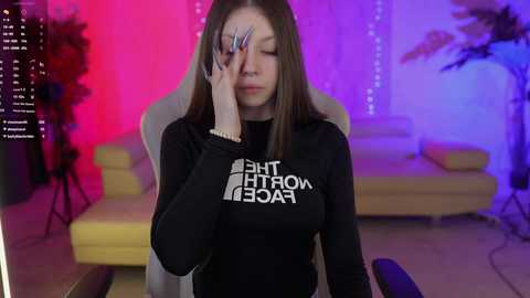 Media: Video of a young woman with straight brown hair, wearing a black sweatshirt with \"THE WORLD IS A GAME\" text, sitting in a yellow gaming chair. The room has purple and pink neon lights, a white couch, and a plant.