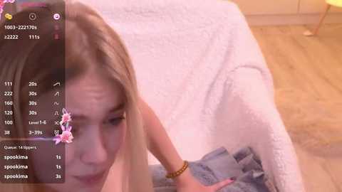 Media: A video of a young blonde woman with long hair, wearing a blue top, lying on a bed with a white quilt, in a cozy room with wooden flooring.