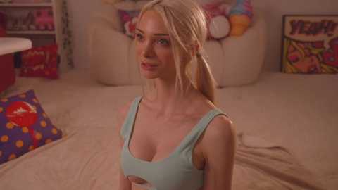 Media: Video of a blonde, fair-skinned woman in a light blue tank top, sitting on a beige bed with colorful pillows and plush toys in the background.