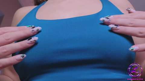 Media: A close-up video of a woman's chest in a tight, blue tank top, with hands placed over her breasts. She has long, manicured nails with intricate, glittery designs. The background is blurred.