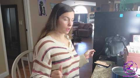 Media: Video of a fair-skinned woman with long brown hair, wearing a striped sweater, giving a thumbs-up in a cozy living room with hardwood floors, a TV, and a brown sofa.