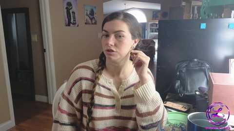 Media: Video of a young woman with light skin, dark hair in braids, wearing a striped sweater, adjusting her earring in a cluttered kitchen.