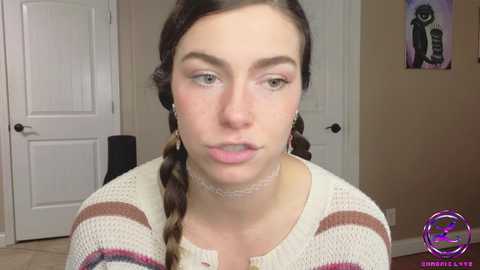 Media: Video of a young woman with fair skin, brown hair in braids, wearing a striped knit sweater, and a choker necklace, indoors with beige walls and a closed white door in the background.