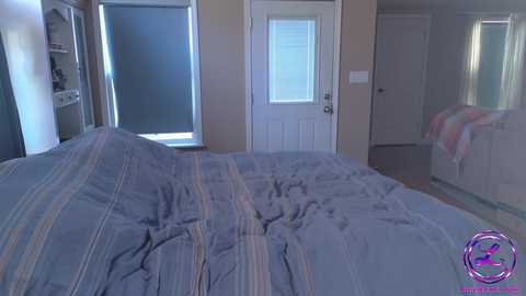 Media: Video of a tidy, modern bedroom with a gray and beige striped bedspread, a white door with blinds, a closet, and a partially visible white dresser.