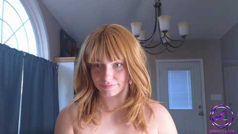 Media: Video of a topless woman with long, straight, auburn hair and fair skin, standing indoors with beige walls, a white door, and a window with a circular glass panel.