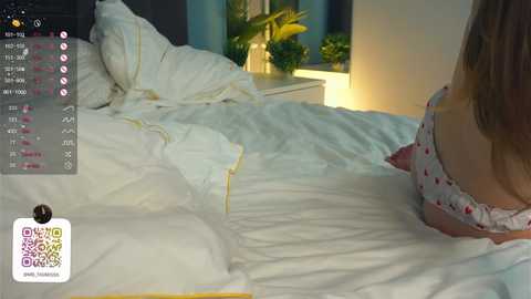 Media: Video of a woman in a pink polka-dot nightgown lying on a white bed with a dark headboard. The room has a potted plant and a nightstand with a lamp.