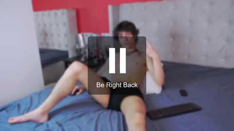 Media: A blurred video of a shirtless man with a slim build and dark hair, wearing black underwear, sitting on a bed with blue sheets, holding a smartphone.