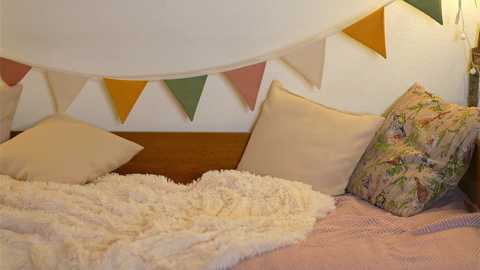 Media: Video of a cozy, pastel-colored bedroom with a fluffy white blanket, beige pillows, and a floral-patterned pillow. Triangular pennant flags hang on a white wall behind.