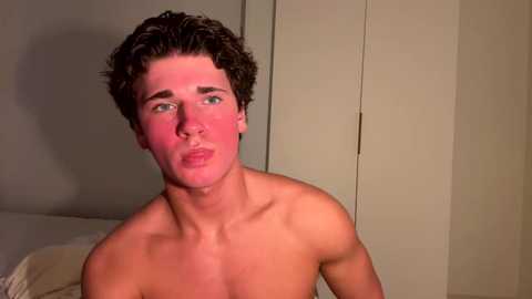 Media: Video of a shirtless young man with curly brown hair, freckles, and fair skin, puckering his lips in a playful expression. Background features a plain, beige wall and a white bed.