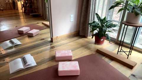 Media: A video of a cozy, light-filled yoga room with wooden floors, a mirror, pink exercise mats, a potted plant, and natural light streaming through large windows.