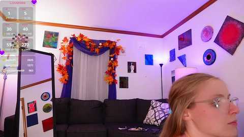 Media: Video of a cozy living room with a woman wearing glasses, a black sofa, orange and purple decor, and various framed artworks.