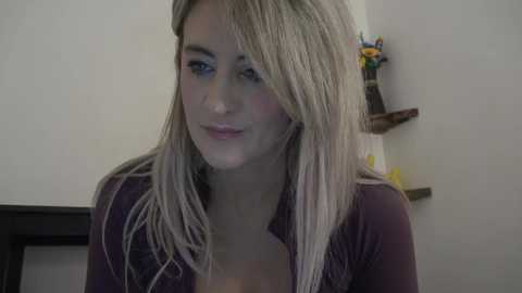 Media: A video of a young, fair-skinned woman with straight, blonde hair, wearing a dark purple top, smiling gently, indoors, with a shelf with toys in the background.