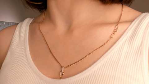 Media: Video of a person with light skin wearing a white ribbed tank top and a delicate gold necklace with a small pendant, blurred background.