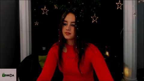 Media: A video of a young Latina woman with long dark hair, wearing a red long-sleeve top, seated indoors with a dimly lit background featuring glowing stars and green foliage.