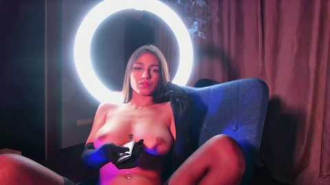 Media: Video of a nude, blonde woman with medium-sized breasts, sitting on a chair, wearing black gloves, with a bright ring light in the background.